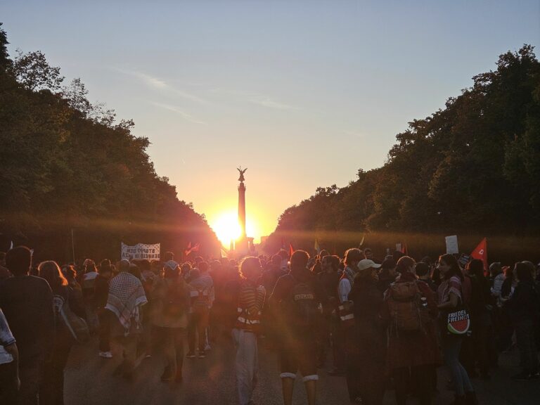 Im Gegenlicht des Sonnununtergangs hinter der Siegessäule in Berlin sieht man Demonstrierende mit Schildern von hinten die Straße des 17. Juni entlanglaufen.